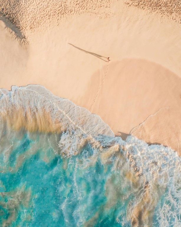 aerial-view-of-beach-with-clear-blue-water-and-single-person-walking-along-shoreline-leaving-footprints