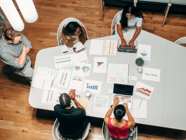 four-people-around-table-with-documents-charts-and-laptop-in-professional-meeting-top-down-view