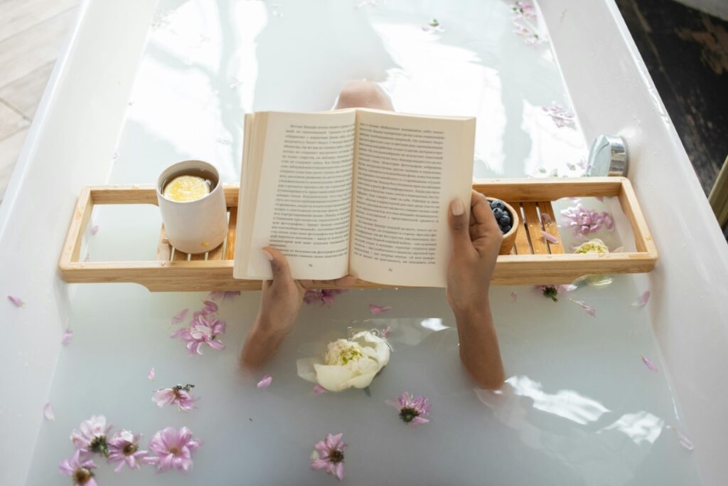 person-in-bathtub-holding-book-with-pink-flowers-and-lit-candle-on-wooden-tray