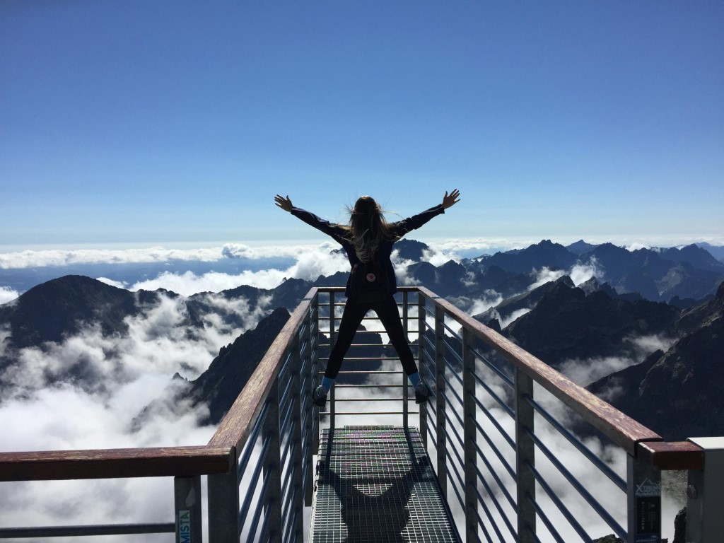 person-standing-on-mountain-walkway-above-clouds-with-arms-outstretched-surrounded-by-mountain-peaks