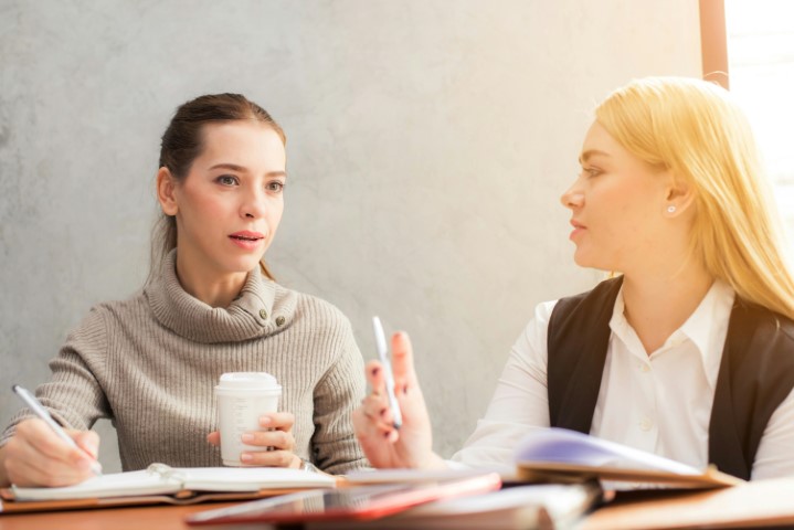 two-people-sitting-at-table-one-writing-other-holding-coffee-cup-faces-obscured